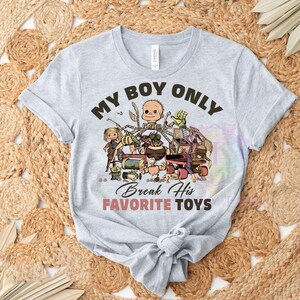 K&ouml;nnte beinhalten: Ein hellgraues Kurzarm-T-Shirt mit einem Grafikdruck aus verschiedenen kaputten Spielzeugen und einem Puppenkopf. Der Text lautet MY BOY ONLY Break His FAVORITE TOYS in schwarzer und rosa Schrift. Das Shirt hat einen Rundhalsausschnitt und einen Knoten.