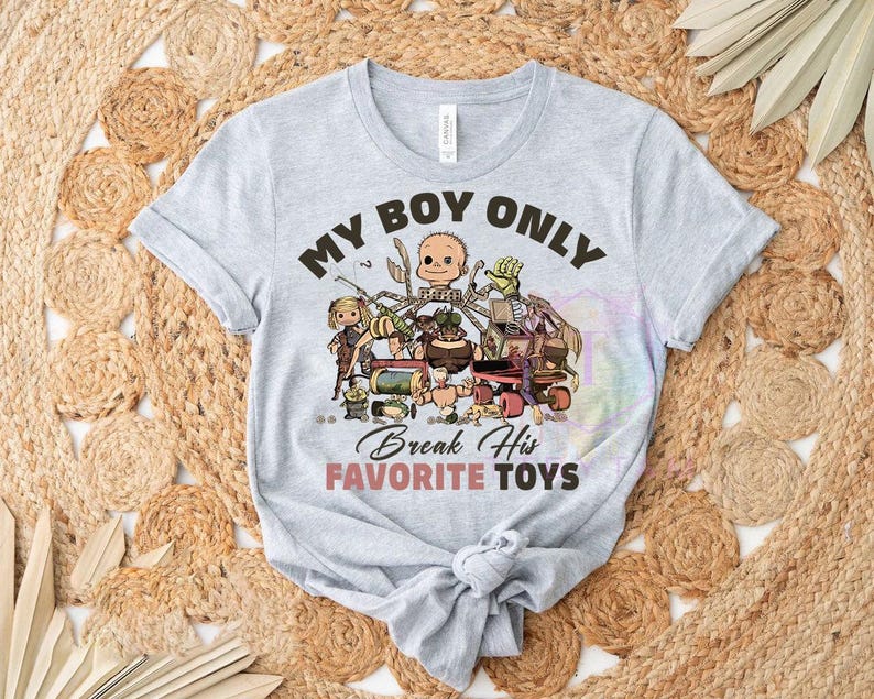 K&ouml;nnte beinhalten: Ein hellgraues Kurzarm-T-Shirt mit einem Grafikdruck aus verschiedenen kaputten Spielzeugen und einem Puppenkopf. Der Text lautet MY BOY ONLY Break His FAVORITE TOYS in schwarzer und rosa Schrift. Das Shirt hat einen Rundhalsausschnitt und einen Knoten.