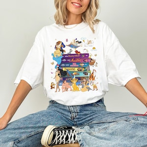 Puede incluir: Camiseta blanca con un dise&ntilde;o de libros apilados de pel&iacute;culas de Disney, incluyendo "La Bella y la Bestia", "Aladdin", "Pocahontas" y "El Rey Le&oacute;n". El dise&ntilde;o presenta personajes de dibujos animados. La camiseta es de corte holgado.