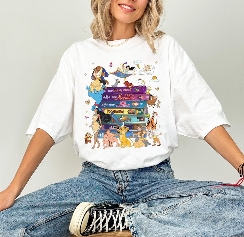 Puede incluir: Camiseta blanca con un dise&ntilde;o de libros apilados de pel&iacute;culas de Disney, incluyendo "La Bella y la Bestia", "Aladdin", "Pocahontas" y "El Rey Le&oacute;n". El dise&ntilde;o presenta personajes de dibujos animados. La camiseta es de corte holgado.