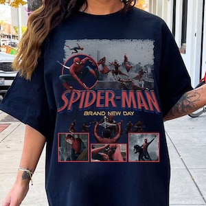May include: Navy blue t-shirt featuring Spider-Man in action poses with the text "SPIDER-MAN BRAND NEW DAY" in red and blue. The design includes multiple panels with Spider-Man illustrations, set against a cityscape backdrop.
