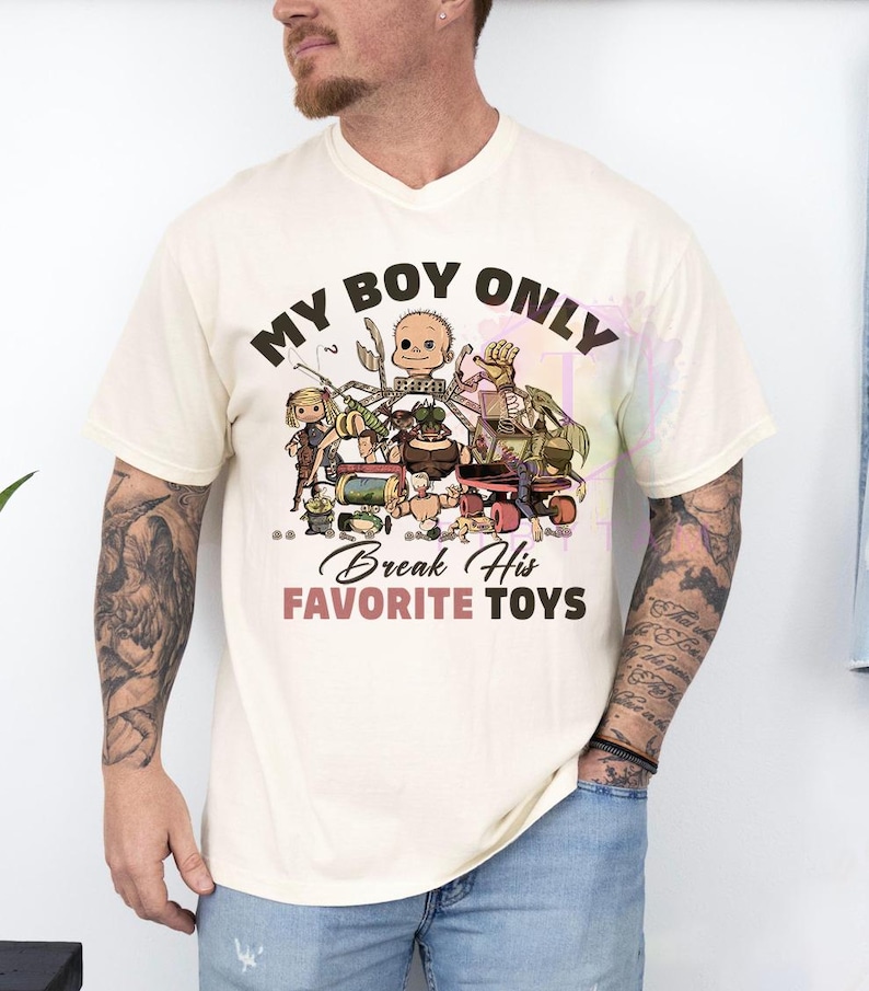 K&ouml;nnte beinhalten: Ein cremefarbenes Kurzarm-T-Shirt mit einem Grafikdruck verschiedener Spielzeuge und eines Puppenkopfes. Der Text MY BOY ONLY steht oben und Break His FAVORITE TOYS unten in schwarzer und rosa Schrift, zentriert auf der Brust.