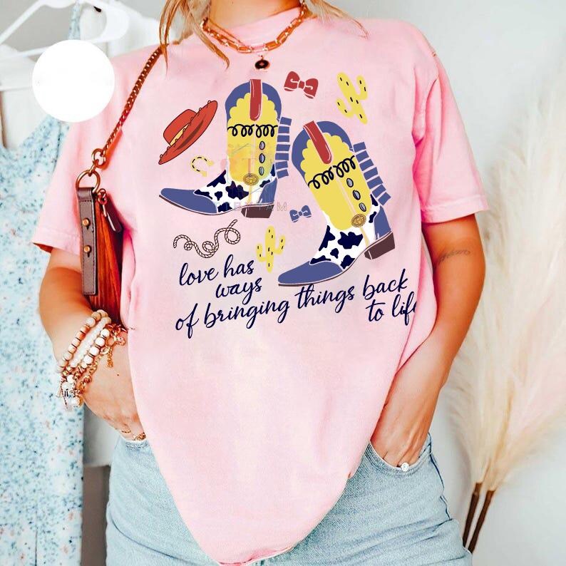 Pu&ograve; includere: Una t-shirt a maniche corte rosa chiaro con una stampa grafica di due stivali da cowboy gialli e bianchi con stampa mucca, un cappello rosso, un cactus e un fiocco. Il testo recita, love has ways of bringing things back to life.
