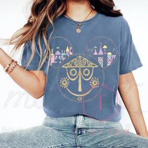 May include: A blue t-shirt featuring a whimsical design. The design incorporates a stylized face and architectural elements within Mickey Mouse ear shapes. The artwork is in shades of gold, pink, purple, and blue, creating a playful and colorful aesthetic.