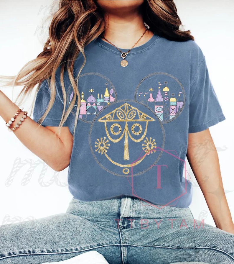 May include: A blue t-shirt featuring a whimsical design. The design incorporates a stylized face and architectural elements within Mickey Mouse ear shapes. The artwork is in shades of gold, pink, purple, and blue, creating a playful and colorful aesthetic.