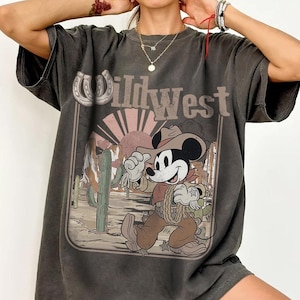 May include: Dark gray t-shirt featuring a graphic of Mickey Mouse dressed as a cowboy in a Wild West scene. The design includes a horseshoe, cactus, and a setting sun. The text "Wild West" is displayed above the graphic.