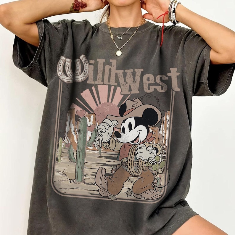 May include: Dark gray t-shirt featuring a graphic of Mickey Mouse dressed as a cowboy in a Wild West scene. The design includes a horseshoe, cactus, and a setting sun. The text "Wild West" is displayed above the graphic.