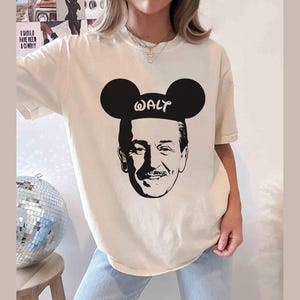 Puede incluir: Camiseta color crema con una silueta negra de Walt Disney con orejas de Mickey Mouse, con la palabra "WALT" impresa en las orejas. La camiseta es de corte holgado.