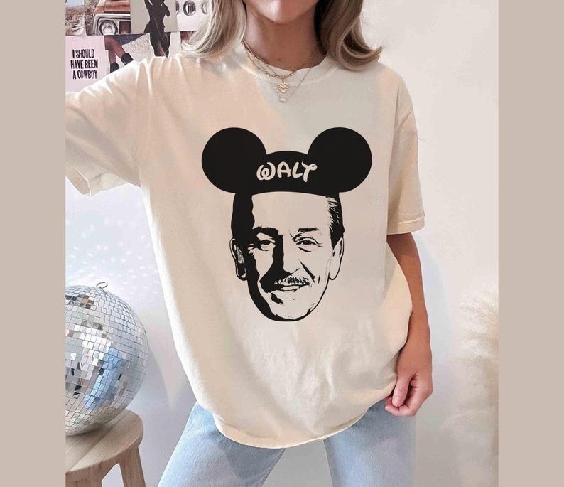 Puede incluir: Camiseta color crema con una silueta negra de Walt Disney con orejas de Mickey Mouse, con la palabra "WALT" impresa en las orejas. La camiseta es de corte holgado.