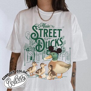Op de afbeelding: Wit comfort color T-shirt met een groene afbeelding van een eend met een Mickey Mouse-hoed, omringd door eendenkuikens en de tekst "Main Street Ducks Co." Het ontwerp bevat een stadsgezicht achtergrond.