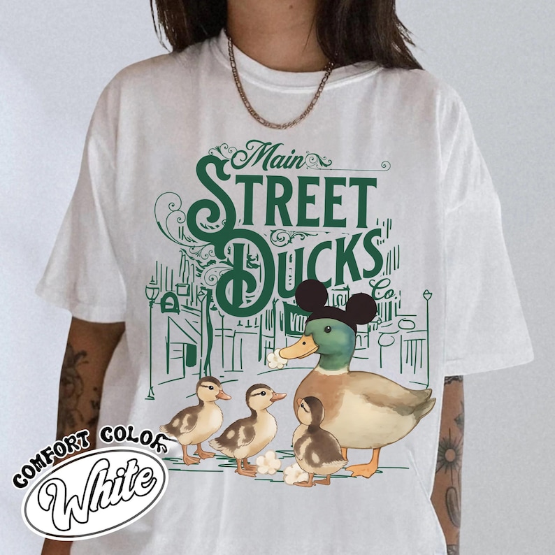 Op de afbeelding: Wit comfort color T-shirt met een groene afbeelding van een eend met een Mickey Mouse-hoed, omringd door eendenkuikens en de tekst "Main Street Ducks Co." Het ontwerp bevat een stadsgezicht achtergrond.