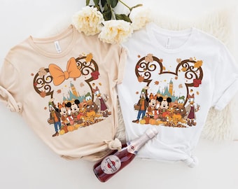 Puede incluir: Dos camisetas de manga corta, una beige y otra blanca, cada una con un gráfico de personajes de Disney que incluye a Mickey, Minnie, Goofy, Donald y Daisy en un entorno otoñal con hojas que caen, calabazas y la silueta de un castillo.