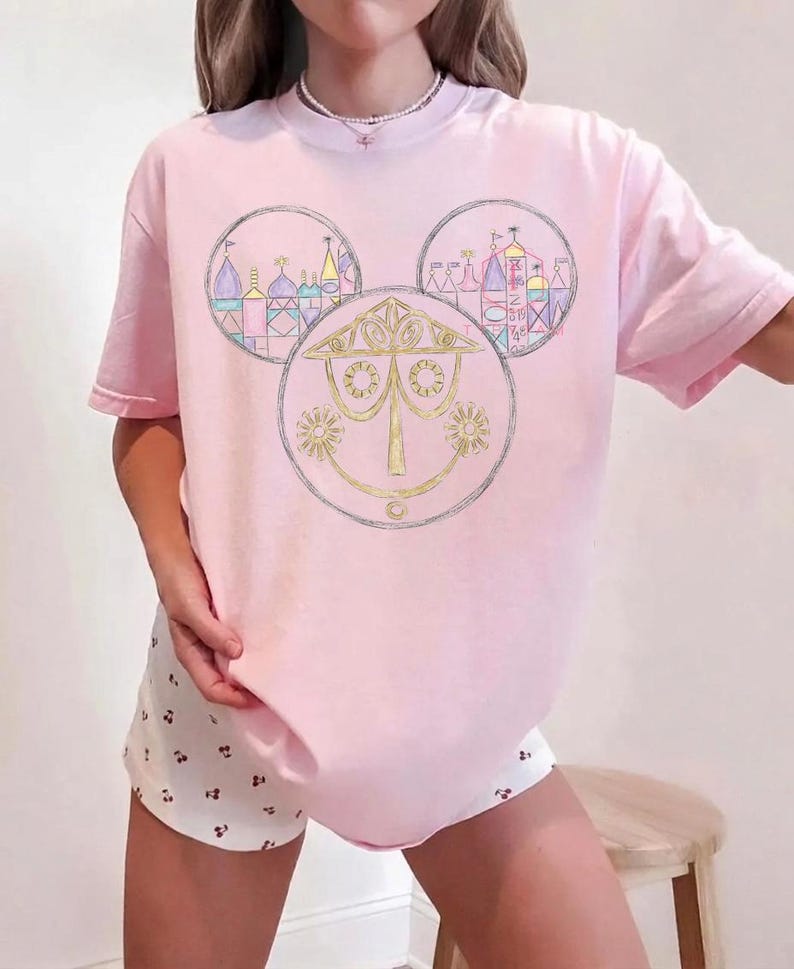 May include: A light pink t-shirt featuring a gold and silver graphic design of a Mickey Mouse head with three circles. Each circle contains a different image of a building. The shirt has short sleeves.