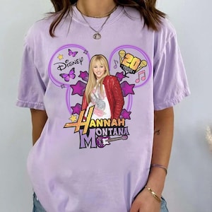 May include: A light purple t-shirt featuring a graphic of Hannah Montana. The design includes the Disney logo, butterflies, stars, and the text "Hannah Montana". The shirt has a relaxed fit.
