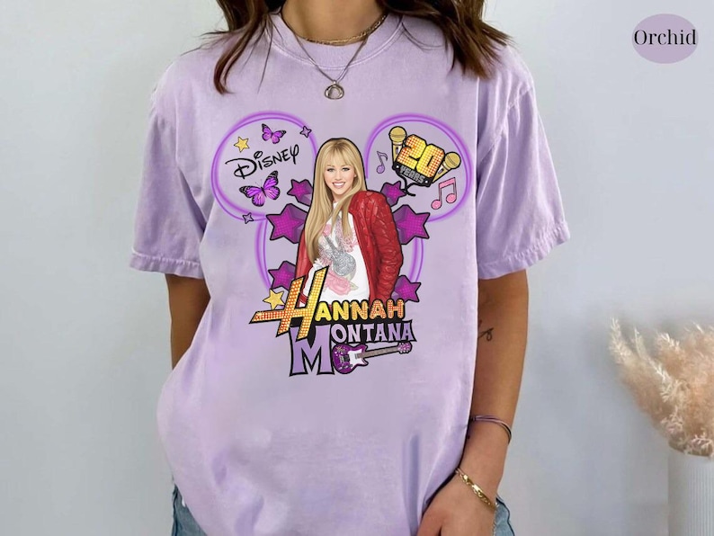 May include: A light purple t-shirt featuring a graphic of Hannah Montana. The design includes the Disney logo, butterflies, stars, and the text "Hannah Montana". The shirt has a relaxed fit.