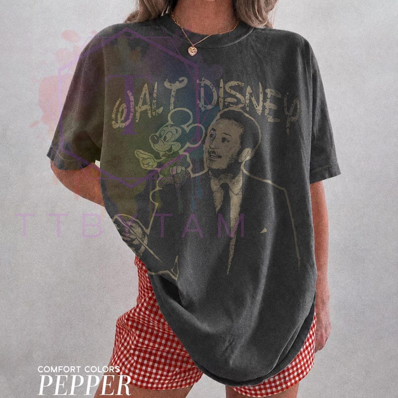 Może przedstawiać: Ciemnoszary t-shirt z kr&oacute;tkim rękawem z grafiką w stylu vintage przedstawiającą Walta Disneya i Myszkę Miki. Napis "WALT DISNEY" jest nadrukowany przetartą czcionką nad postaciami. Koszulka ma luźny kr&oacute;j i wygląda na lekko zużytą.