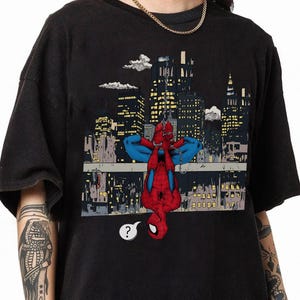 May include: Black t-shirt featuring a comic book illustration of Spider-Man hanging upside down against a cityscape backdrop. The superhero is depicted in his classic red and blue suit, with a question mark speech bubble.