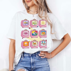 May include: White t-shirt featuring a grid of pink retro televisions, each displaying a different cartoon or show title. The televisions have a decorative design and are arranged in three rows. The shirt is worn by a person.