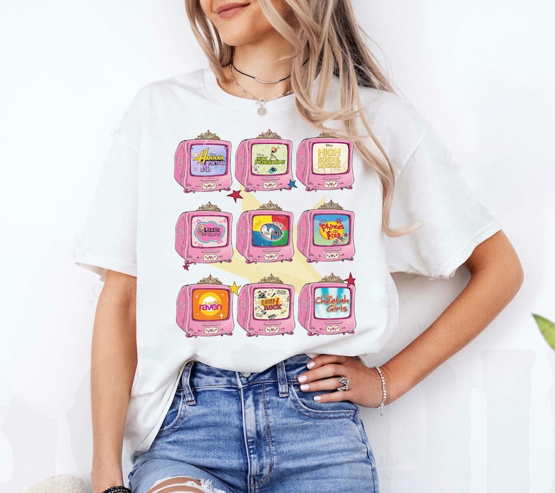 May include: White t-shirt featuring a grid of pink retro televisions, each displaying a different cartoon or show title. The televisions have a decorative design and are arranged in three rows. The shirt is worn by a person.
