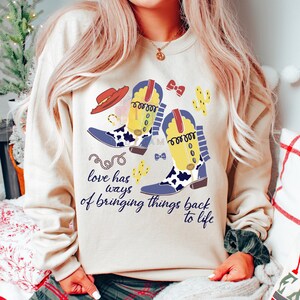 Pu&ograve; includere: Una felpa beige a girocollo con una stampa grafica di due stivali da cowboy gialli e blu con dettagli maculati, un cappello rosso, cactus e una corda. Il testo "love has ways of bringing things back to life" &egrave; stampato in corsivo blu scuro.