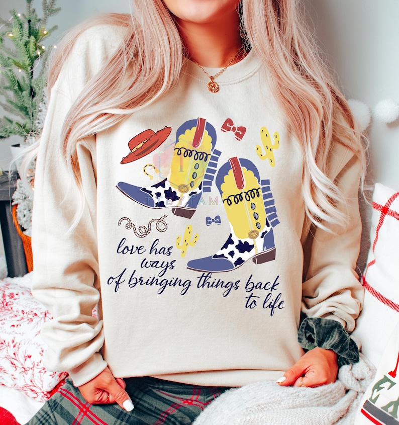 Pu&ograve; includere: Una felpa beige a girocollo con una stampa grafica di due stivali da cowboy gialli e blu con dettagli maculati, un cappello rosso, cactus e una corda. Il testo "love has ways of bringing things back to life" &egrave; stampato in corsivo blu scuro.