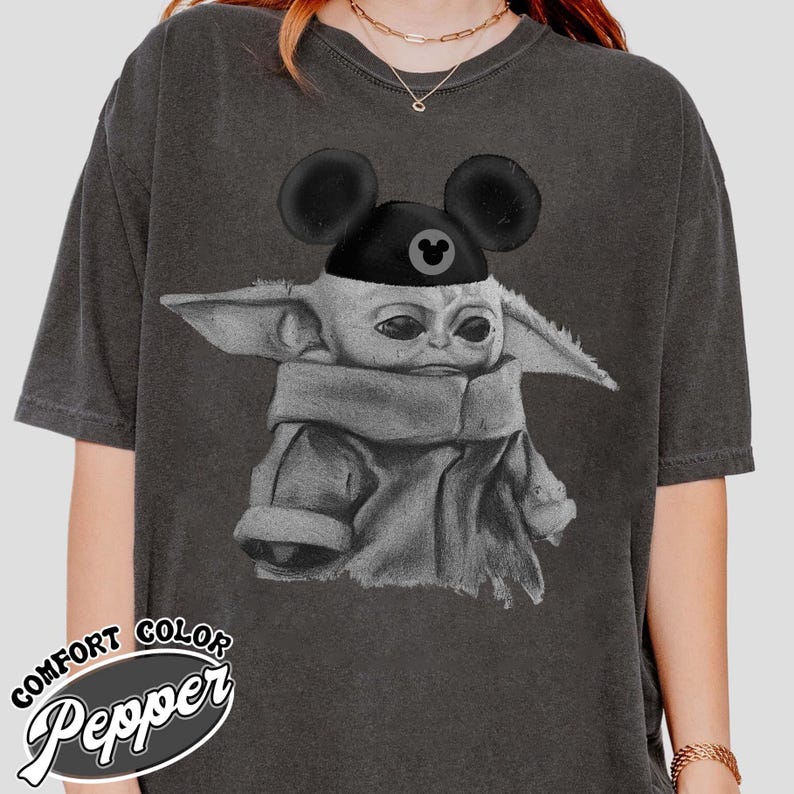 May include: A charcoal gray short-sleeve t-shirt featuring a graphic illustration of a small creature with large ears wearing a black cap with mouse ears. The shirt has a distressed, washed-out appearance and includes a logo in the corner.