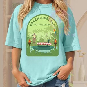 Puede incluir: Camiseta azul claro con un gr&aacute;fico verde que dice "ADVENTURELAND - NATIONAL PARK". El gr&aacute;fico representa una escena de la jungla con un barco, figuras, palmeras y un loro. Antorchas a ambos lados del barco.