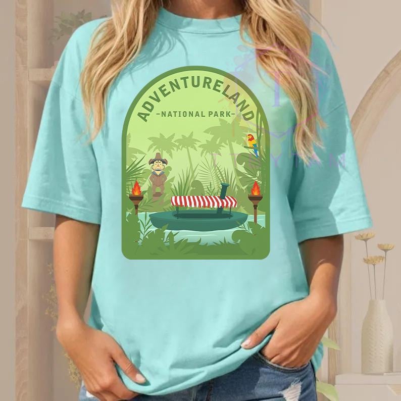 Puede incluir: Camiseta azul claro con un gr&aacute;fico verde que dice "ADVENTURELAND - NATIONAL PARK". El gr&aacute;fico representa una escena de la jungla con un barco, figuras, palmeras y un loro. Antorchas a ambos lados del barco.