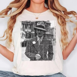 May include: A cream-coloured t-shirt with a black and white vintage image of a man in a suit and hat. The image has the word "Walt" overlaid in white. The man is wearing a suit, tie, and a hat with a flower on his lapel.