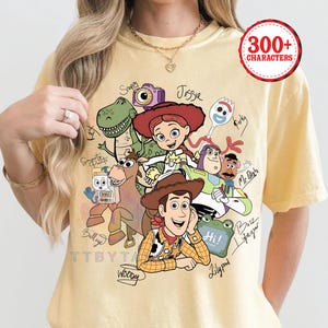 May include: A pale yellow t-shirt featuring a graphic print of Toy Story characters including Woody, Jessie, Buzz Lightyear, Rex, Bullseye, Forky, and Mr. Potato Head. The design includes handwritten-style names next to each character and the text 300+ CHARACTERS.