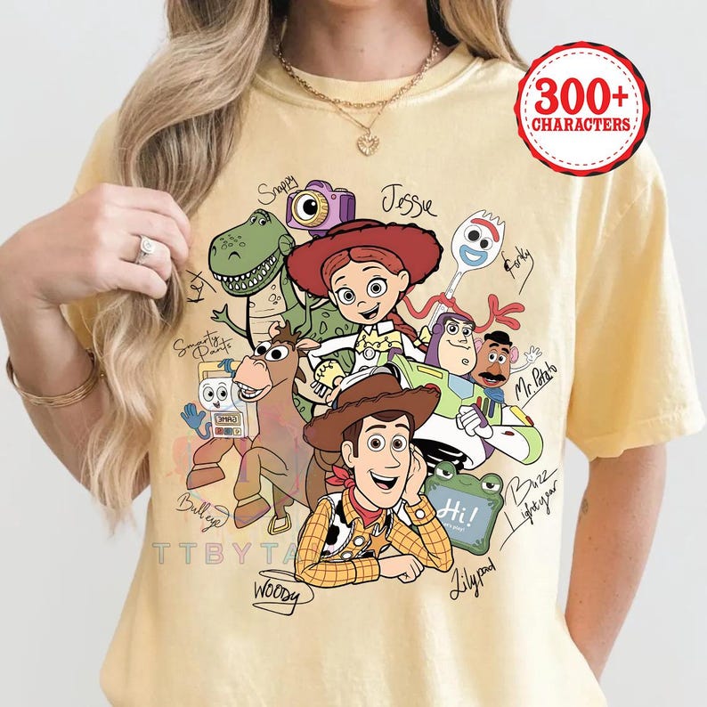 May include: A pale yellow t-shirt featuring a graphic print of Toy Story characters including Woody, Jessie, Buzz Lightyear, Rex, Bullseye, Forky, and Mr. Potato Head. The design includes handwritten-style names next to each character and the text 300+ CHARACTERS.