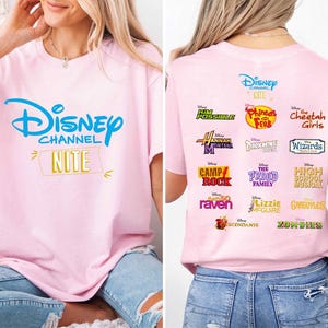 May include: Pink t-shirt featuring the Disney Channel logo in blue, with "NITE" in a yellow banner. The back of the shirt displays logos from various Disney Channel shows, including "Kim Possible," "Phineas and Ferb," and "The Cheetah Girls."