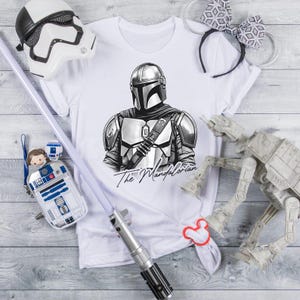 K&ouml;nnte beinhalten: Wei&szlig;es T-Shirt mit einer Schwarz-Wei&szlig;-Illustration von The Mandalorian. Das Bild enth&auml;lt einen Stormtrooper-Helm, ein Lichtschwert, eine R2-D2-Tasche, ein Paar M&auml;useohren und ein AT-AT-Modell. Der Text "The Mandalorian" steht darunter.