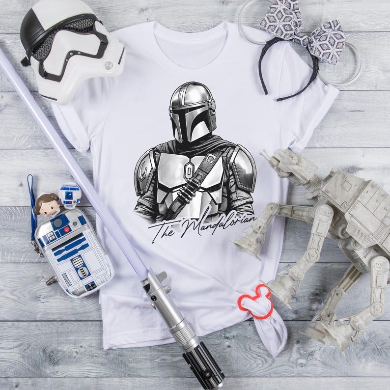 K&ouml;nnte beinhalten: Wei&szlig;es T-Shirt mit einer Schwarz-Wei&szlig;-Illustration von The Mandalorian. Das Bild enth&auml;lt einen Stormtrooper-Helm, ein Lichtschwert, eine R2-D2-Tasche, ein Paar M&auml;useohren und ein AT-AT-Modell. Der Text "The Mandalorian" steht darunter.
