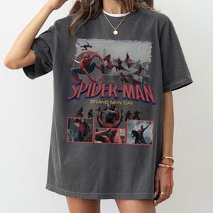 May include: Dark gray t-shirt featuring the Spider-Man logo and the words "BRAND NEW DAY" in red and blue. The shirt has a graphic of Spider-Man in action, with additional smaller images below the logo.