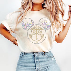 May include: Cream-colored t-shirt featuring a Mickey Mouse-shaped design. The design includes three circles: two with colorful castle illustrations and one with a gold-colored, stylized face. The shirt is made of soft material.