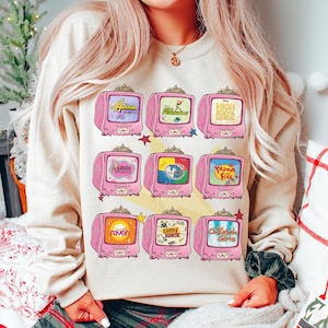 May include: Beige sweatshirt featuring a pink television graphic with various show titles, including "Hannah Montana," "High School Musical," and "Lizzie McGuire." The graphic has a retro aesthetic with star accents.