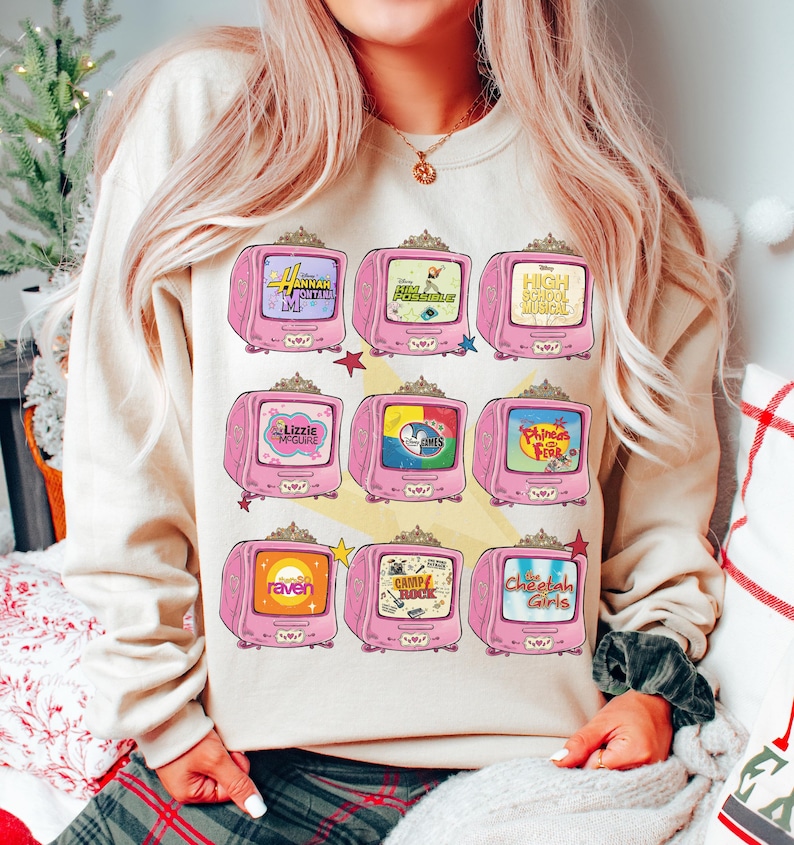 May include: Beige sweatshirt featuring a pink television graphic with various show titles, including "Hannah Montana," "High School Musical," and "Lizzie McGuire." The graphic has a retro aesthetic with star accents.