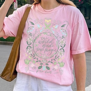 Pu&ograve; includere: T-shirt rosa chiaro con un design floreale e la scritta "the world needs your magic". Il design include fiori, foglie e le parole "faith" e "trust". Le parole "pixie dust" sono nella parte inferiore del design.