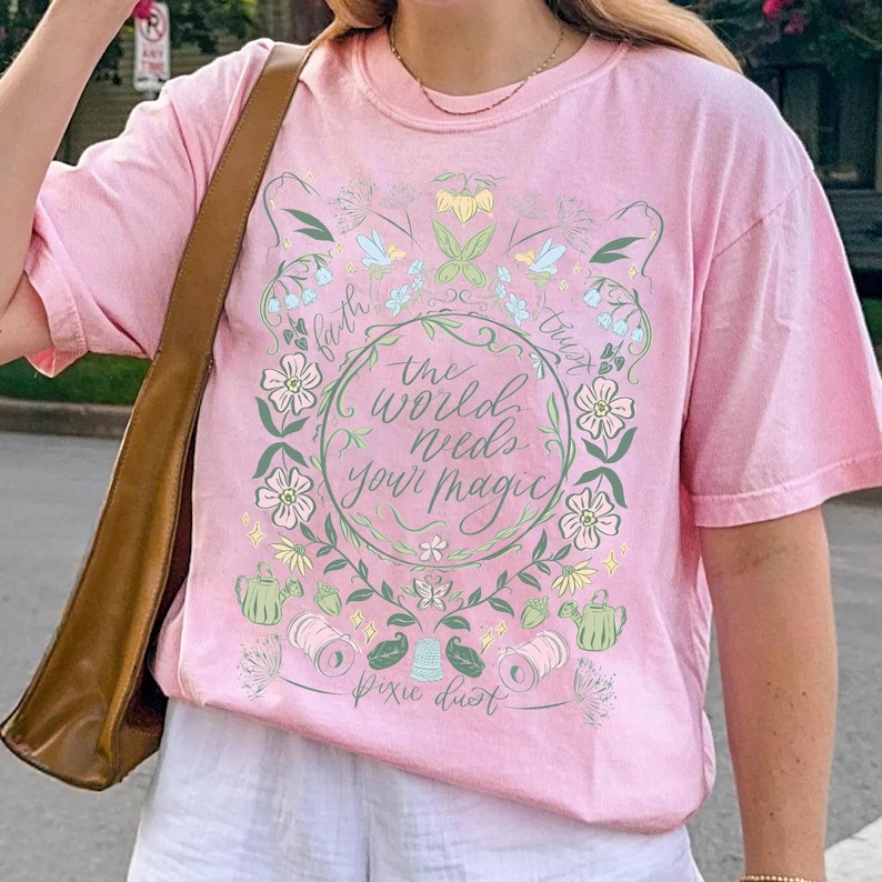 Pu&ograve; includere: T-shirt rosa chiaro con un design floreale e la scritta "the world needs your magic". Il design include fiori, foglie e le parole "faith" e "trust". Le parole "pixie dust" sono nella parte inferiore del design.