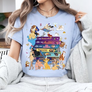 Puede incluir: Camiseta azul claro con un dise&ntilde;o de cintas VHS coloridas con t&iacute;tulos y personajes de pel&iacute;culas de Disney. El dise&ntilde;o incluye personajes de La Bella y la Bestia, Aladdin, Pocahontas y El Rey Le&oacute;n.