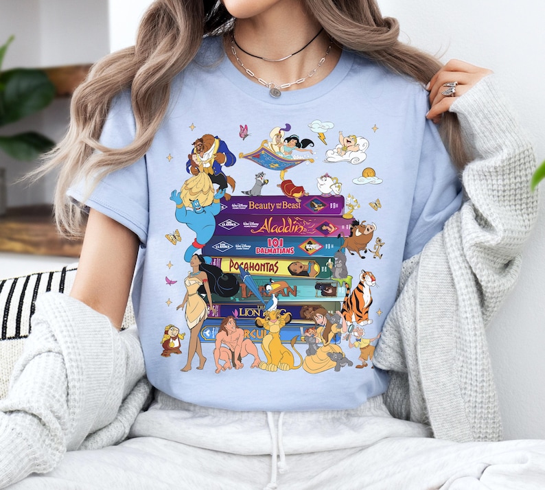 Puede incluir: Camiseta azul claro con un dise&ntilde;o de cintas VHS coloridas con t&iacute;tulos y personajes de pel&iacute;culas de Disney. El dise&ntilde;o incluye personajes de La Bella y la Bestia, Aladdin, Pocahontas y El Rey Le&oacute;n.