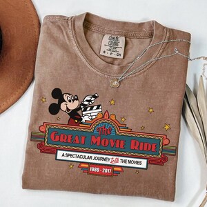 Puede incluir: Camiseta marr&oacute;n con Mickey Mouse sosteniendo una claqueta de cine. El texto en la camiseta dice "The Great Movie Ride" con los a&ntilde;os 1989-2017. Un collar plateado con un colgante est&aacute; sobre la camiseta.