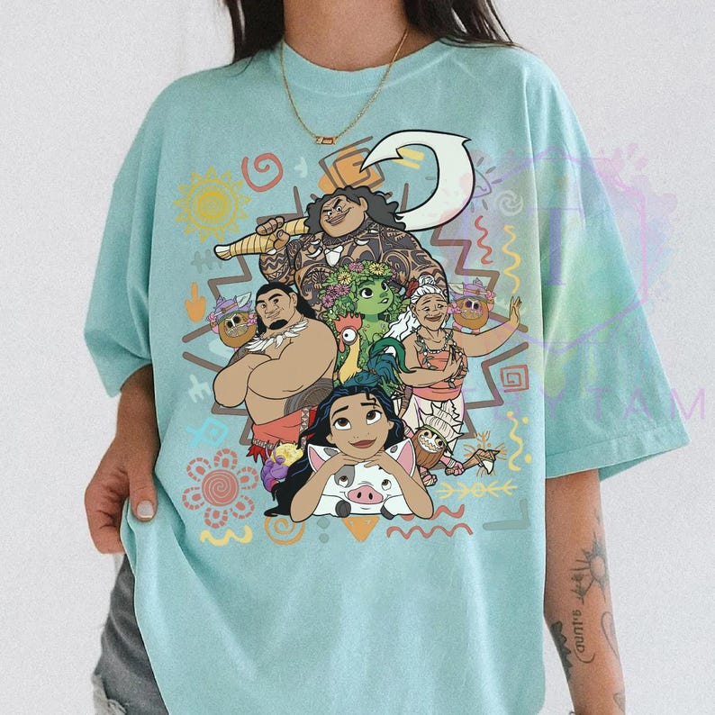 May include: A light blue t-shirt featuring a colorful graphic of characters from a popular animated movie. The design includes a variety of characters, a large hook, and decorative tribal patterns. The shirt is a comfortable, casual style.