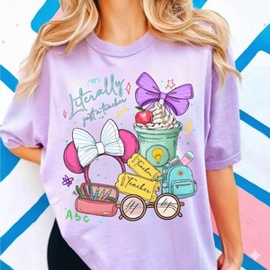 May include: A light purple t-shirt with the text "Literally just a teacher" and a graphic of school-related items. The graphic includes a coffee cup, apple, bow, glasses, pencils, and a backpack.