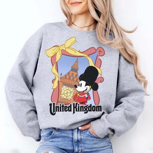 May include: A light gray sweatshirt featuring a cartoon image of Mickey Mouse in a British guard uniform, standing in front of Big Ben. The image is framed with a pink border and a yellow bow. The words "United Kingdom" are printed below.