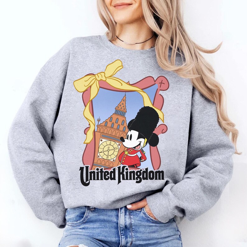 May include: A light gray sweatshirt featuring a cartoon image of Mickey Mouse in a British guard uniform, standing in front of Big Ben. The image is framed with a pink border and a yellow bow. The words "United Kingdom" are printed below.