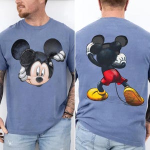 May include: A blue t-shirt featuring a Mickey Mouse graphic on the front and back. The front graphic shows Mickey's face, while the back shows Mickey in a dynamic pose. The design includes black, red, yellow, and white colors.