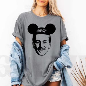 Puede incluir: Camiseta gris con un gr&aacute;fico negro de Walt Disney con orejas de Mickey Mouse y la palabra "WALT". La camiseta tiene un corte holgado y se combina con una chaqueta vaquera y pantalones cortos.