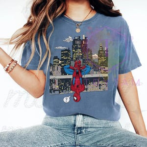 May include: Blue t-shirt featuring a comic-style illustration of Spider-Man hanging upside down in front of a city skyline. The graphic includes a question mark speech bubble. The shirt is a solid blue color.